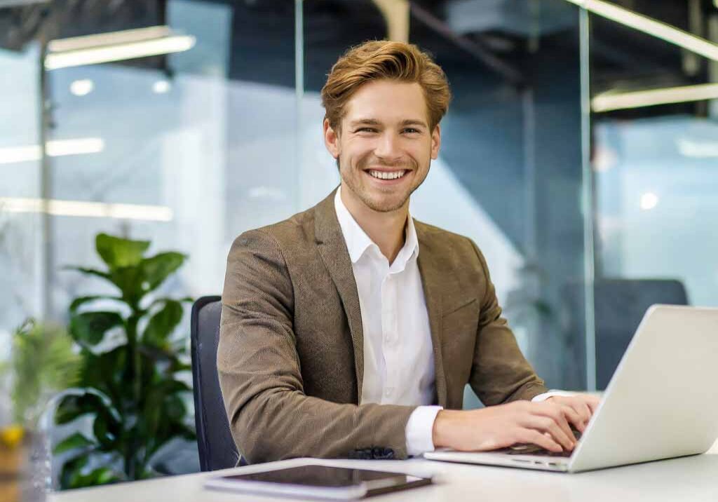 Firefly a man working on a laptop sitting at a desk in a modern office. He has a big smile looking f Firefly a man working on a laptop sitting at a desk in a modern office. He has a big smile looking f
