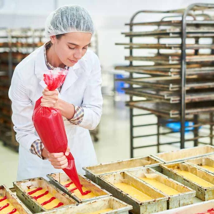 Confectionery factory employee using pastry bag Confectionery factory worker in white coat pouring red cream from pastry bag.